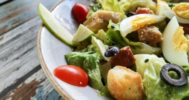 A close-up of a Caesar salad featuring fresh lettuce, eggs, croutons, and olives on a rustic table.